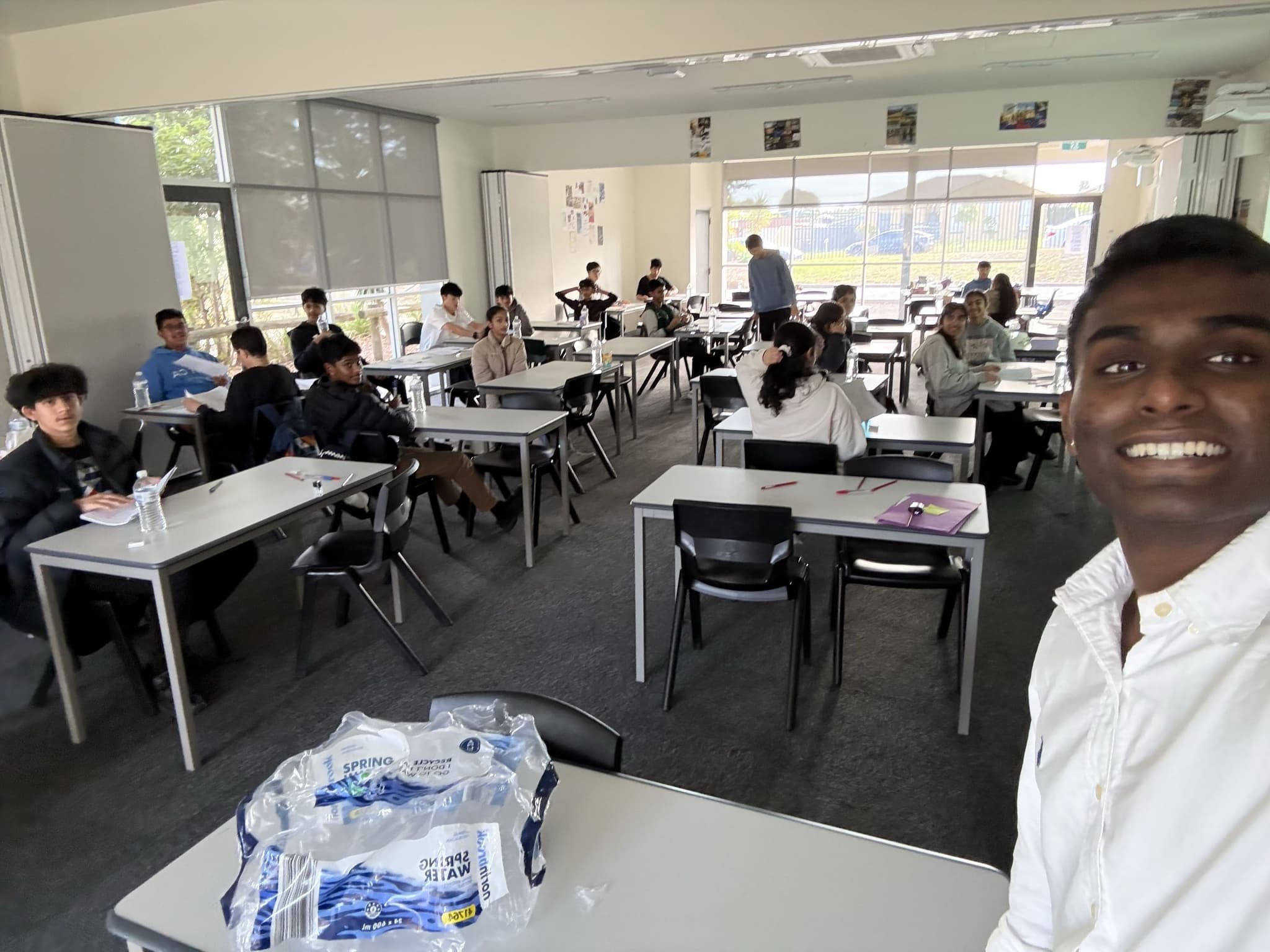 Dev smiling in a selfie at the front of a busy weekend class, students working at desks behind him.